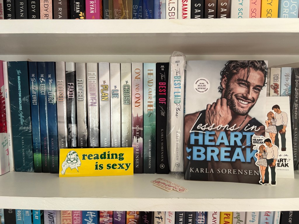 A shelf filled with various romance novels, prominently featuring the book 'Lessons in Heartbreak' by Karla Sorensen, alongside a playful sign stating 'reading is sexy'.