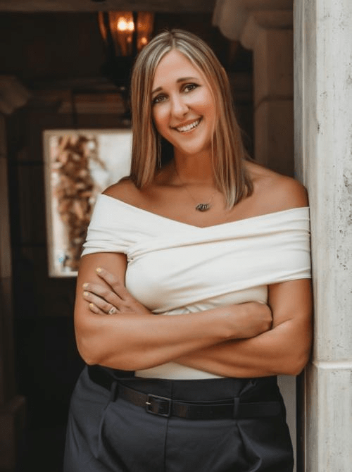 A smiling woman with long blonde hair, wearing an off-shoulder white top and black pants, stands beside a stone wall in a well-lit space, with her arms crossed.