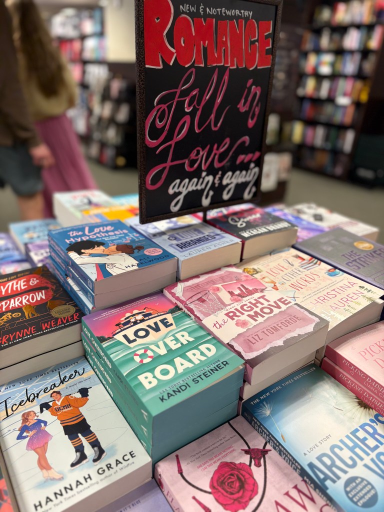 Display of romance novels stacked on a table with a sign that reads 'New & Noteworthy Romance - Fall in Love again & again.'